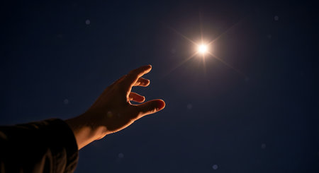A person's hand is seen reaching up towards a single, bright, glowing star or light in a clear, dark night sky. The image conveys a sense of hope, aspiration, desire, or reaching for a goal.の素材