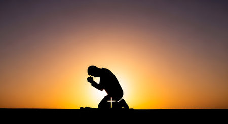 A silhouette of a man kneeling in prayer on a hilltop against a vibrant orange and purple sunset. A small white cross is visible on his leg, symbolizing Christian faith, worship, and hope.の素材