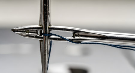 An extreme macro shot showing a blue thread passing through the eye of a sewing needle, which itself is passed through the eye of a second needle. This image illustrates concepts of precision, connection, difficulty, and intricate detail.の素材