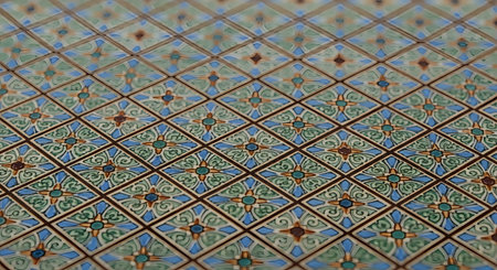 A close-up, angled shot of an intricate tile pattern with a repeating geometric and floral design. The tiles feature blue, green, and orange colors within a diamond-shaped grid, showcasing detailed ceramic or paper craftsmanship.の素材