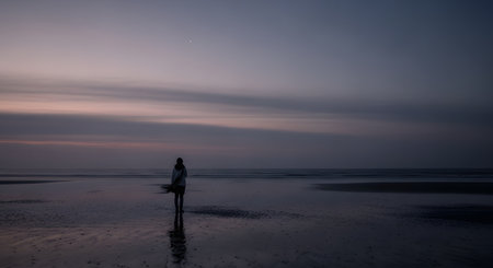 A lone person is silhouetted on a wide, wet, reflective beach, looking out at the calm ocean during twilight. The sky is a soft gradient of purple and pink, creating a serene, moody, and contemplative atmosphere.の素材