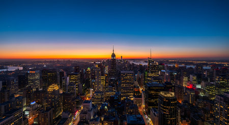 A stunning wide-angle aerial view of the Manhattan skyline at twilight. The sky transitions from a warm orange and yellow glow at the horizon to a deep blue, while the city buildings, including the iconic Empire State Building, are illuminated with lights.の素材