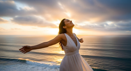 A happy woman in a white dress stands on a cliff overlooking the ocean at sunset, with her arms outstretched. She has her eyes closed and a peaceful smile, embracing freedom, joy, and nature.の素材
