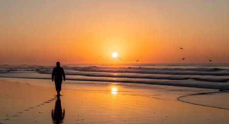 A silhouette of a man walking alone on a wet sandy beach during a vibrant orange sunrise or sunset. His reflection is visible on the sand, and birds are flying in the sky over the calm ocean waves. This image evokes peace, solitude, and contemplation.の素材