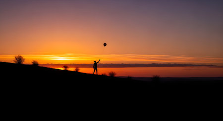 A striking silhouette of a person standing on a dark hill, holding a single balloon up against a vibrant, colorful sunset. The sky transitions from bright orange and yellow at the horizon to deep purple and blue. The image evokes feelings of freedom, hope, loneliness, or celebration.の素材