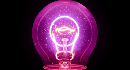 A macro close-up of a clear glass light bulb illuminated against a black background. The filament and internal structure are glowing with a vibrant, energetic pink and purple light, creating a bright circular halo effect inside the bulb.の素材