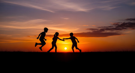 A wide silhouette of three children, two boys and a girl, playing and running in a grassy field against a vibrant sunset. The sun is a bright orange orb on the horizon, set in a dramatic sky of orange, purple, and blue. This image evokes feelings of childhood, joy, freedom, friendship, and nostalgia.の素材