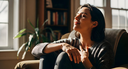 A serene woman sits in a comfortable armchair at home,basking in a warm patch of sunlight streaming from a window. With her eyes closed and a gentle smile,she holds a hand to her chest,embodying mindfulness,gratitude,and relaxation.の素材