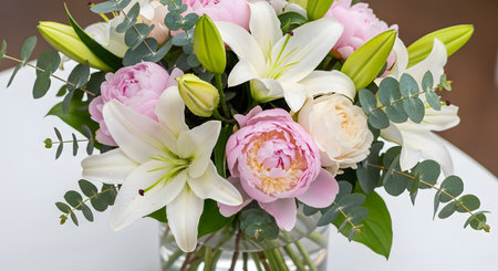 A beautiful and elegant flower bouquet in a clear glass vase, featuring white lilies, pink peonies, and light pink roses, accented with eucalyptus leaves. The fresh arrangement is set on a white table, perfect for a gift, celebration, or home decor.の素材