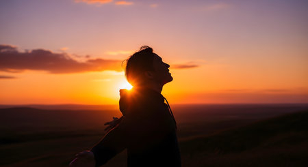A silhouette of a person standing on a hill with arms outstretched, embracing a stunning, colorful sunset. The sky is a warm gradient of orange, yellow, and purple, and the person's head is tilted back in a gesture of freedom, gratitude, or worship. The scene conveys a sense of peace, hope, and connection with nature.の素材