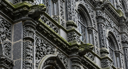 A low-angle shot of a historic building's ornate facade, featuring two detailed stone gargoyles. The intricate gothic architecture, covered in moss, showcases arched windows and complex carvings, evoking a sense of history and mystery.の素材