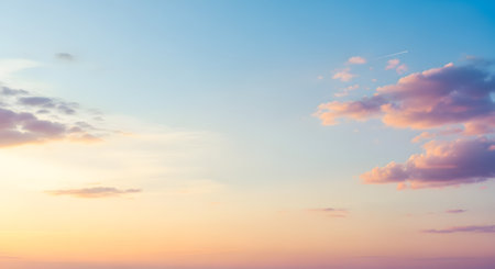 A serene and beautiful pastel sky at dusk or dawn, with a soft gradient from orange and yellow at the horizon to a calm blue above. A few fluffy pinkish clouds are scattered across the sky, with a faint trail from an airplane in the distance, creating a peaceful and dreamy background.の素材