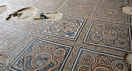 An ancient, intricate mosaic floor, likely Roman or Byzantine, is partially uncovered during an archaeological excavation. The detailed geometric and floral patterns are visible, with sections still covered in dirt and excavation tools like brushes lying nearby.の素材
