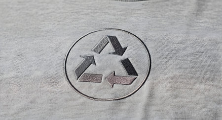 A detailed close-up shot of a recycling symbol embroidered onto a grey fabric t-shirt. The universal symbol with three chasing arrows is stitched in shades of grey and pink, representing environmental consciousness and sustainable fashion. The texture of the cotton fabric is clearly visible.の素材