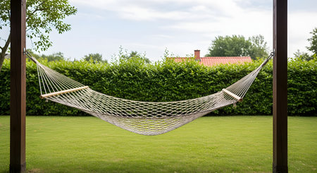 An empty rope hammock is strung between two wooden posts in a lush green backyard. The manicured lawn and a tall green hedge in the background create a peaceful and relaxing scene, perfect for a summer day.の素材