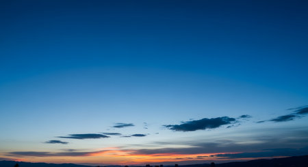 A stunning twilight sky with a deep blue gradient transitioning to warm orange and pink hues at the horizon after sunset. A dark silhouette of a city or mountains rests at the bottom under a few wispy clouds.の素材