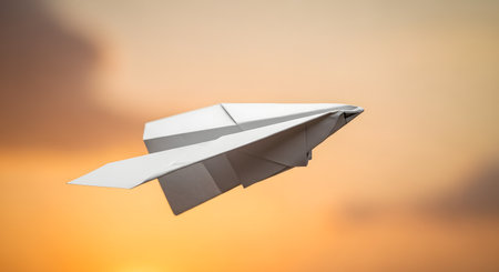 A white paper airplane is captured in mid-flight, soaring against a beautifully blurred, warm orange and yellow sunset sky. The image symbolizes freedom, travel, dreams, childhood, and imagination.の素材