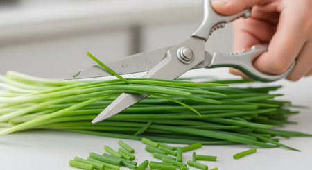 A close-up shot of a person's hand using stainless steel kitchen scissors to cut a bunch of fresh, vibrant green chives. Some chopped pieces are already on the white cutting surface, illustrating food preparation and cooking with fresh herbs. The background is clean and bright, focusing on the culinary action.の素材