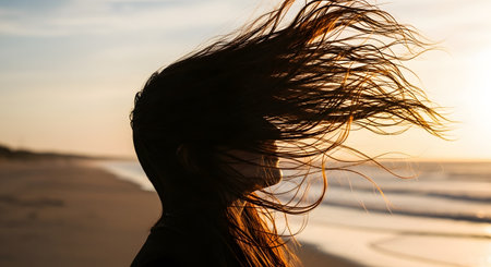 A silhouette of a woman's head in profile, with her long hair blowing dramatically in the wind. She is on a beach at sunset, and the warm, golden sunlight highlights her hair against the blurred ocean background. This image evokes freedom, nature, and serenity.の素材