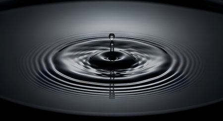 A single drop of water hits a still, dark surface, creating a perfect splash and a series of concentric ripples that expand outwards. The monochrome image symbolizes impact, cause and effect, and tranquility.の素材
