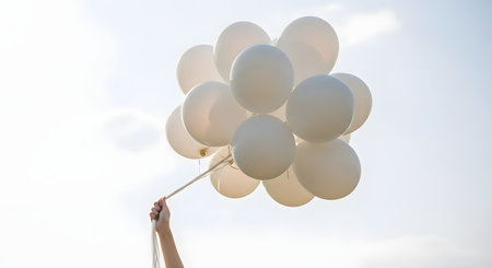 A person's arm and hand are seen from below, holding the strings of a large bunch of white balloons against a bright, pale sky. The image evokes feelings of celebration, freedom, peace, or letting go.の素材
