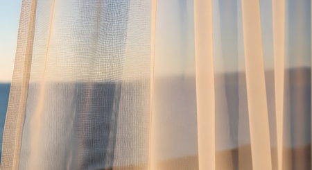 Warm, golden morning sunlight gently filters through a delicate, sheer curtain, creating a soft and peaceful ambiance. The fine texture of the fabric is visible, and the background is a soft-focus view of a calm sea or sky. The image evokes tranquility, relaxation, and a gentle start to the day.の素材