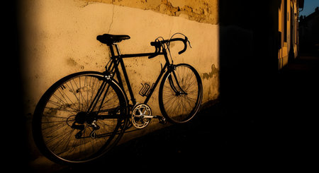 A black road bicycle is parked, leaning against a textured, weathered wall bathed in the warm, dramatic light of the golden hour. The low sun casts long shadows, creating a tranquil and nostalgic urban scene.の素材