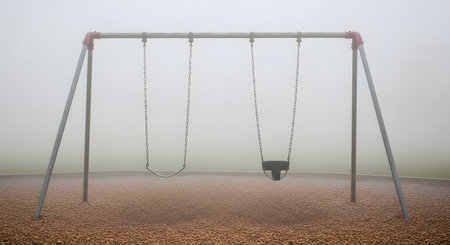 An empty playground swing set, featuring one regular swing and one baby swing, stands on a wood chip surface. The entire scene is enveloped in thick, white fog, creating a mysterious, eerie, and lonely atmosphere.の素材