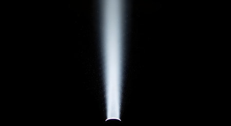 A single, bright beam of white light shines down from a spotlight, cutting through a black background. Tiny dust particles are illuminated within the ray, creating a focused, atmospheric effect. This image represents illumination, focus, and discovery.の素材