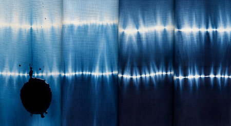 A beautiful background of blue and white Japanese shibori or tie-dye fabric, showing a repeating pattern from the dyeing technique. A large, dark ink blot or paint splatter sits on the left side, adding an element of artistic imperfection or a creative accident to the textile.の素材