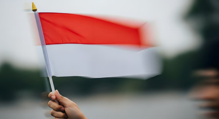 A person's hand holds and waves a small Indonesian flag, with intentional motion blur creating a dynamic sense of movement and energy. The image conveys patriotism, celebration, national pride, and support for Indonesia.の素材