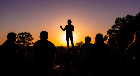 A silhouette of a person standing and speaking to a group of people sitting on the ground. The scene is set on a hilltop at sunset, with the sky in warm orange and purple hues. This image represents community, storytelling, or leadership.の素材