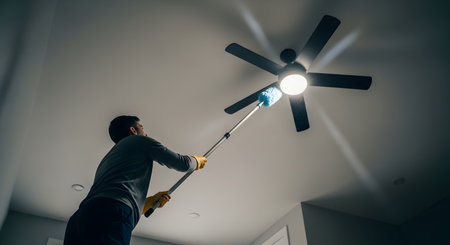 A low-angle view of a man using an extendable blue duster to clean the blades of a ceiling fan. Dust particles are visible in the air, illuminated by the fan's light, as he performs this household chore.の素材