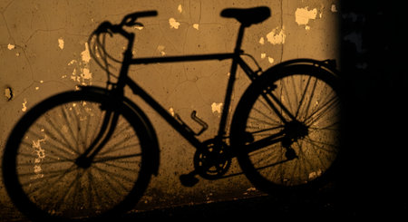 The dark shadow of a bicycle is cast onto a rustic, peeling yellow wall by the warm light of a low sun. The silhouette clearly defines the bike's frame, wheels, and handlebars, creating a nostalgic or urban scene.の素材