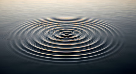 Perfect concentric circles or ripples spread outward from a central point on the surface of calm, dark water. The image, reflecting the soft light of the sky, evokes feelings of peace, zen, meditation, and the concept of impact or a chain reaction.の素材