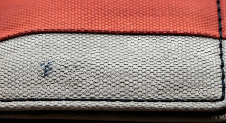 An extreme macro, close-up shot of a wallet or bag made from two-tone canvas fabric (red-orange and light grey/beige). The focus is on the detailed weave and texture of the material, with dark stitching along the seams. This image is an ideal textile background.の素材