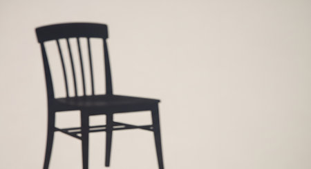 The dark, blurry shadow of a classic wooden spindled chair is cast onto a plain, beige-colored wall. The minimalist, abstract image creates a sense of presence, absence, or solitude. This image represents concepts of waiting, emptiness, or memory.の素材