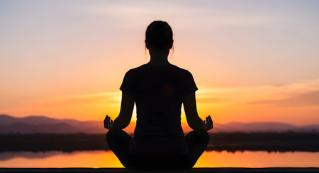The silhouette of a woman seen from behind, meditating in a yoga lotus pose on a wooden deck. She faces a serene lake and mountains, bathed in the warm, orange glow of a beautiful sunset. This image evokes peace, mindfulness, wellness, and harmony with nature.の素材