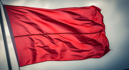 A large, vibrant red flag billows in the wind, attached to a flagpole, against a dramatic, overcast sky. The image can symbolize a warning, danger (a red flag), or a signal.の素材