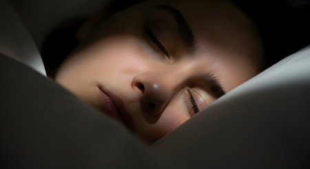A close-up, intimate shot of a young woman's face as she sleeps peacefully. Her eyes are closed, and her face is partially in shadow, nestled in white pillows and blankets, conveying rest and tranquility.の素材