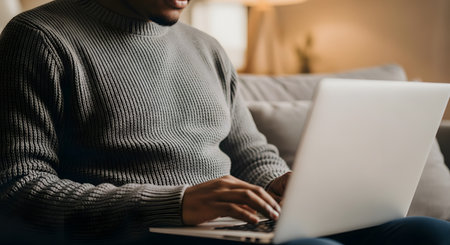 A close-up shot of an African American man wearing a grey knitted sweater, sitting on a couch and typing on a laptop. The man is focused on his work, representing concepts like working from home, remote work, or casual web browsing.の素材