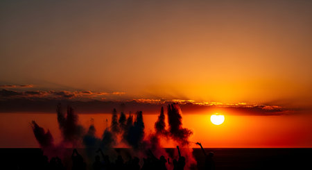 A silhouette of a crowd of people at a beach festival, set against a stunning orange sunset. Plumes of dark (possibly colored) smoke rise from the crowd, suggesting smoke bombs or pyrotechnics. The atmosphere is energetic, festive, and celebratory.の素材