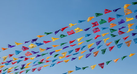 Multiple strings of colorful 'papel picado' banners are strung diagonally against a clear, bright blue sky. The festive paper flags, in various colors like red, yellow, blue, and green, are cut with intricate designs, symbolizing a celebration or festival.の素材