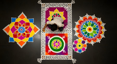 An overhead, top-down view of a person creating intricate and colorful Rangoli patterns on a dark floor. The vibrant designs, made with colored powders, are for a Hindu festival like Diwali or Onam, representing celebration, tradition, and art.の素材