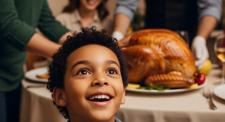A close-up shot of a young boy with curly hair, looking up with a wide, joyful smile of anticipation. In the blurred background, adults are placing a large, roasted turkey on a set dinner table, indicating a holiday feast like Thanksgiving or Christmas.の素材