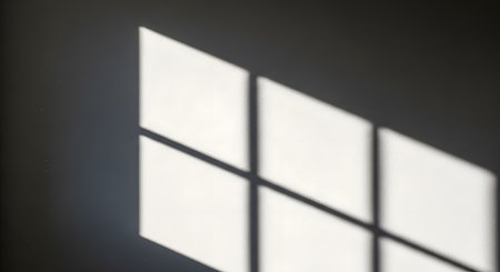 Sunlight streams through a window, casting a distinct shadow of the window pane grid onto a textured wall. The play of light and shadow creates a minimalist and abstract geometric pattern. The image evokes a sense of quiet, calm, and time passing.の素材