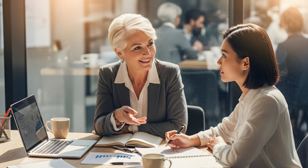 A senior businesswoman with white hair mentors a younger Asian colleague in a bright, modern office. They are sitting at a desk with a laptop displaying charts, smiling and engaged in a productive discussion. The younger woman is taking notes, illustrating concepts of mentorship, collaboration, and professional development.の素材