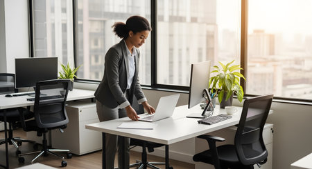 A professional businesswoman in a grey suit stands at her desk in a modern, high-rise office with large windows overlooking a city. She is opening her laptop to begin her workday, representing ambition, career, and corporate life.の素材
