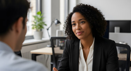 A professional African American businesswoman attentively listens to a male colleague during a meeting in a modern office. She is dressed in a black blazer, conveying confidence and engagement in their discussion. The setting suggests a job interview, consultation, or business negotiation.の素材