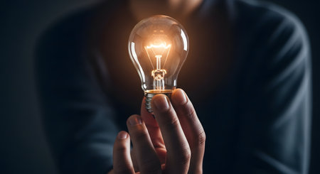 A person in a dark shirt is holding up a classic incandescent lightbulb which is glowing brightly. The lightbulb represents an idea, innovation, creativity, or inspiration.の素材